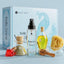 Katari Belle Voyaj - complete Mediterranean ritual set featuring a light blue branded box, a roseau facial toner spray bottle, a small glass jar with a cork lid and green paste, a clear bottle of yellow oil, a red heart-shaped item, cinnamon sticks, and a yellow sponge on a light blue background.