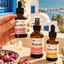 Hand holding Katari Rosehip Oil beside Roseau and Hoba Oil on a Mediterranean terrace with dried rosebuds