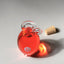 an opened hand blown glass bottle of Granate, Katari pomegranate seed oil, with a cork on a light grey background with a light reflection through the oil
