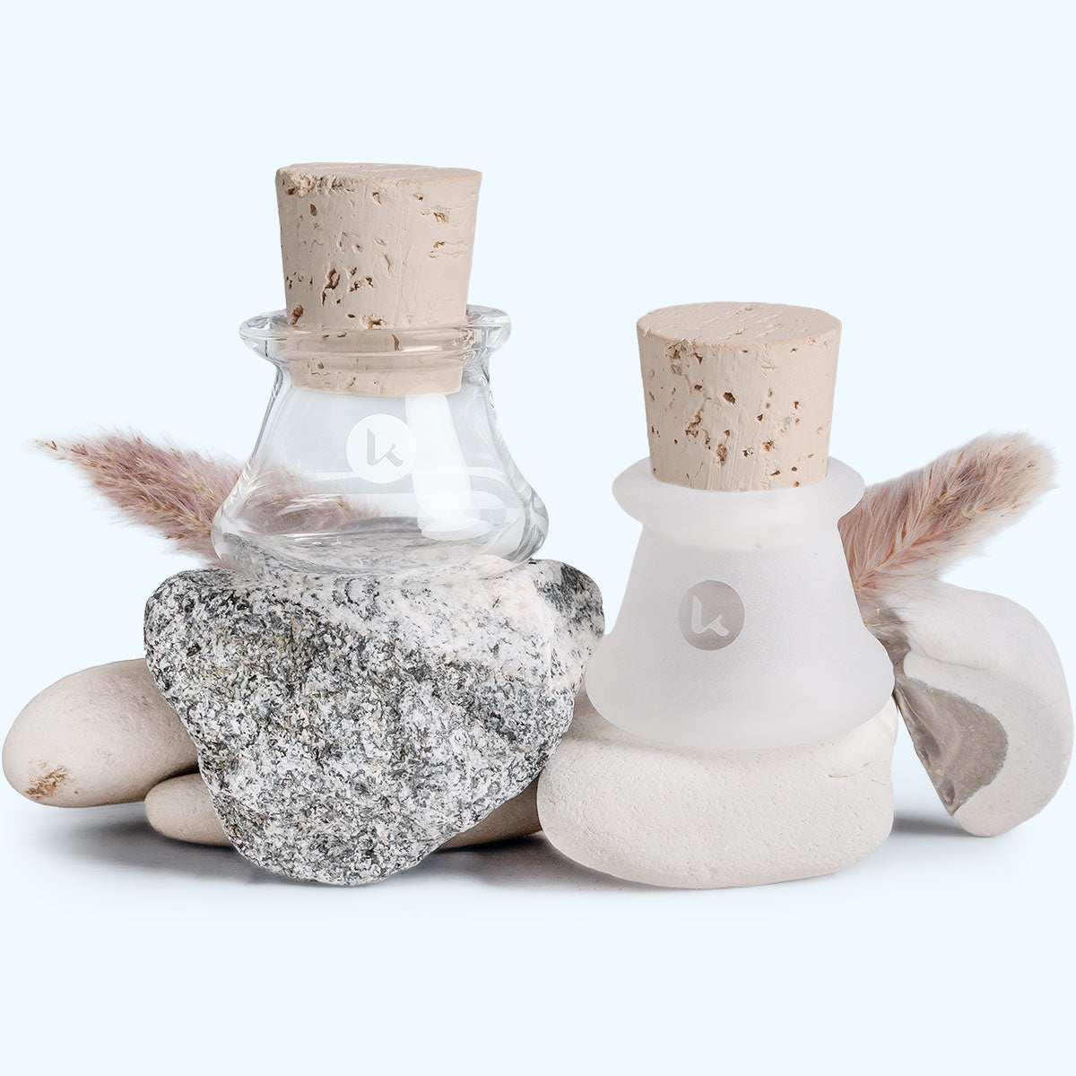 Two hand blown glass jars with cork lids and on a white background set with grey and white rocks and dried grass
