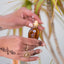 Water splashing on a hand-blown glass amphora of Katari Black Seed Oil, a pure cold-pressed source of thymoquinone (TQ), held by hands with henna art.