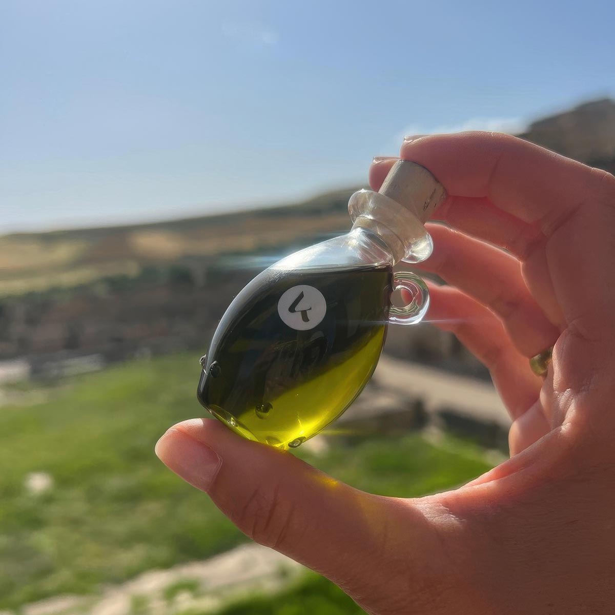 Hand holding a hand-blown glass amphora of Katari Henna Oil for hair and lash growth against a sunny Mediterranean landscape.