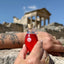 Hand holding a hand-blown glass amphora of Katari Rosehip Oil, rich in Vitamins A and C, with ancient Roman ruins in the background.