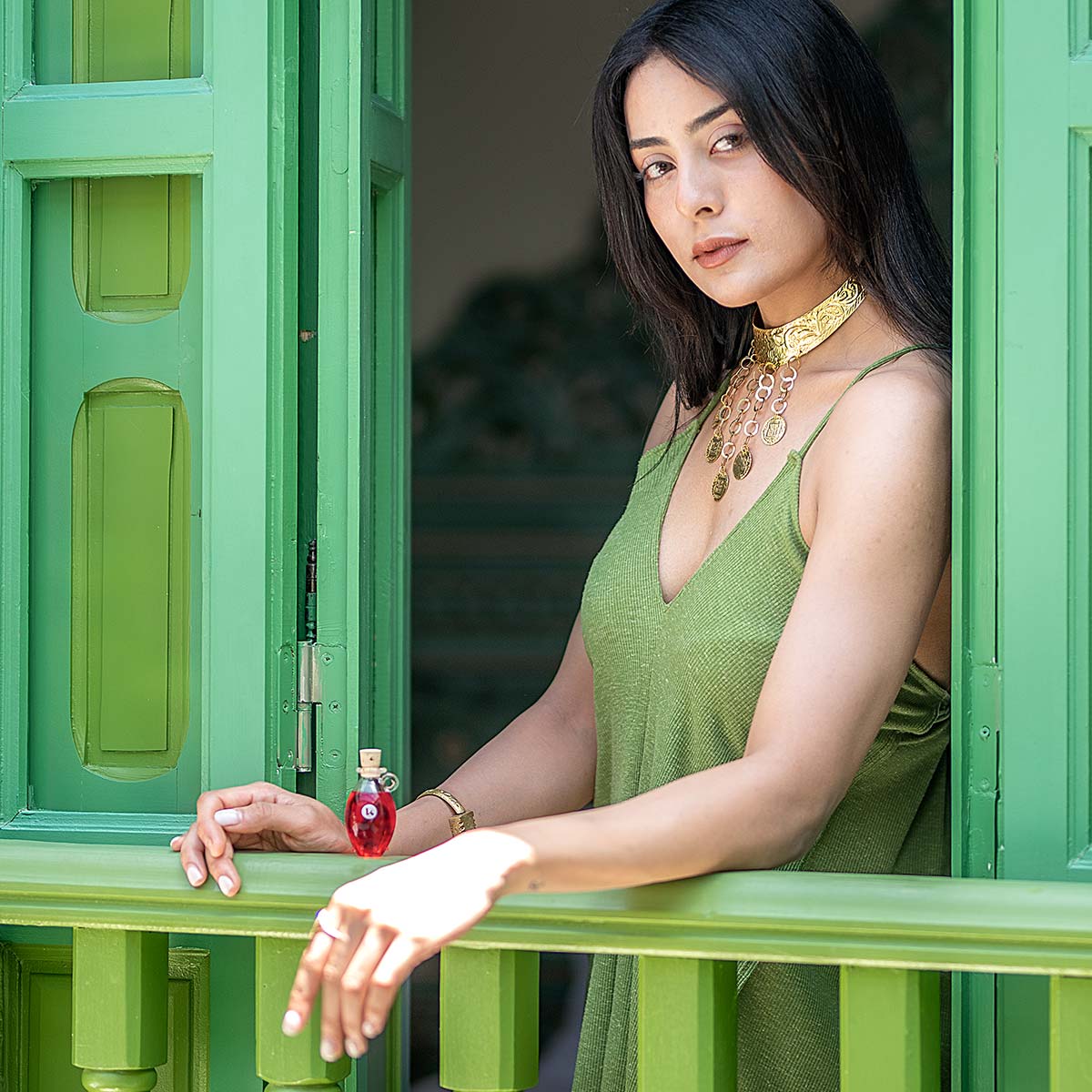 A hand-blown glass amphora of Katari Rosehip Oil, rich in vitamins A, C, and Omegas, on a green balcony with a woman in Mediterranean style.
